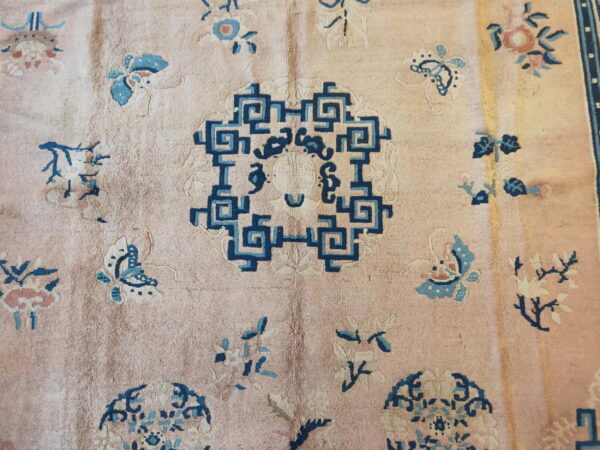 A muted pink rug features a navy geometric key pattern medallion surrounded by scattered blue floral and butterfly motifs.