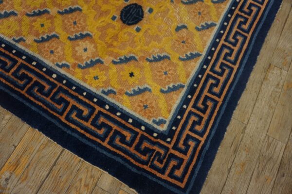 Yellow floral rug corner featuring a navy blue meander border rests on rustic light brown wood flooring.