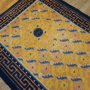 A yellow and peach rug with a repeating floral pattern and a navy Greek key border sits on wood planks.