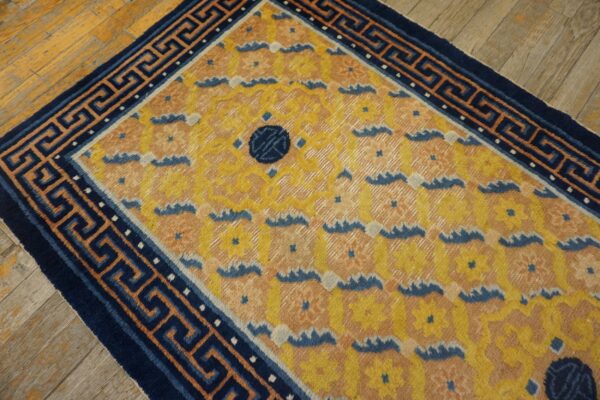 A yellow and peach rug with a repeating floral pattern and a navy greek key border sits on wood planks.