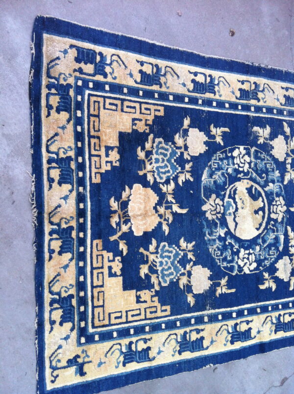 Blue rug with cream floral patterns and intricate geometric key borders on a gray concrete floor.