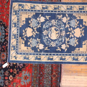 A blue and beige floral rug is centered over red and navy patterned rugs on wood plank flooring.