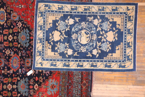 A blue and beige floral rug is centered over red and navy patterned rugs on wood plank flooring.