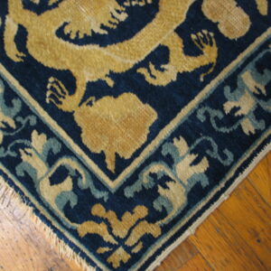 Navy, gold, and teal rug corner with stylized motifs and fringe rests on distressed wood flooring.