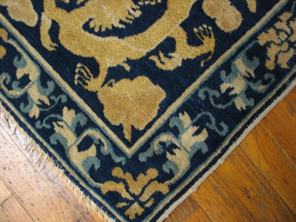 Navy, gold, and teal rug corner with stylized motifs and fringe rests on distressed wood flooring.