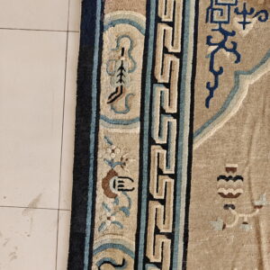 High-pile tan rug border featuring navy, light blue, and beige key patterns rests on white tiled floor.