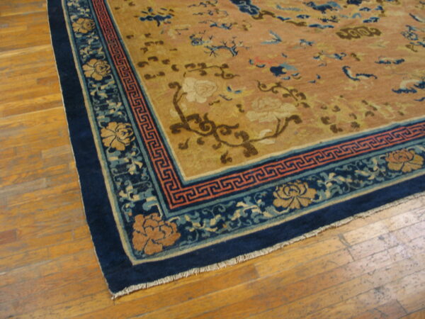 Tan field rug with scattered floral and blue motifs, framed by a dark blue ornamental border on wood flooring.
