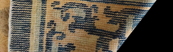 Detail of a low-pile rug featuring a stepped geometric pattern in dark blue, tan, and light green.