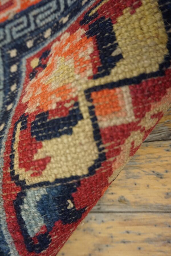 A close-up of a curled rug shows geometric patterns in red, navy, orange, and beige over distressed wood planks.