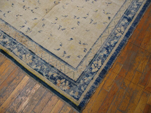 Low-pile beige rug with blue abstract field patterns and a wide ornamental blue border on wood flooring.