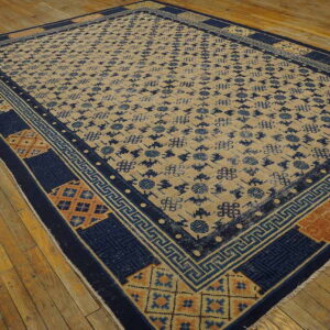 Tan rug with dark blue geometric repeat pattern and intricate borders rests on distressed wood flooring.