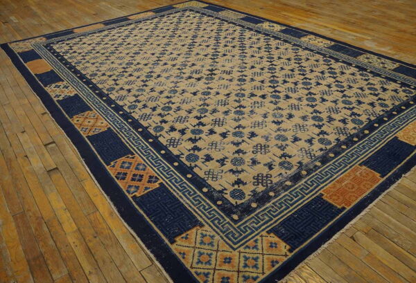 Tan rug with dark blue geometric repeat pattern and intricate borders rests on distressed wood flooring.