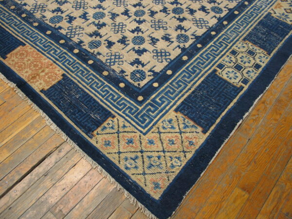 Tan rug with scattered blue motifs and a detailed blue and rust corner border rests on worn amber floorboards.