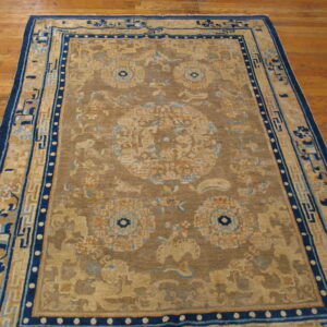 Brown and tan rug featuring stylized blue and beige medallion and wide geometric borders rests on a hardwood floor.
