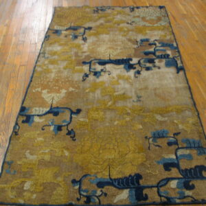 Low-pile, rectangular rug featuring gold, tan, and dark blue abstract designs on hardwood flooring.