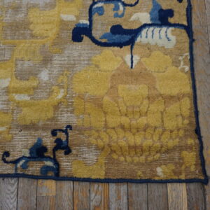 Section of a golden-toned rug with navy blue patterns rests on distressed wooden flooring.