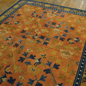 Low pile orange rug with sprawling blue and gold floral patterns and a dark blue border on hardwood flooring.