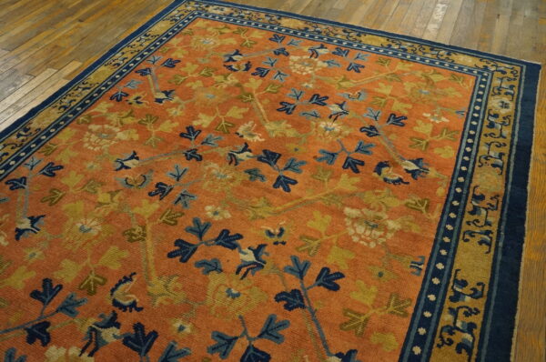 Low pile orange rug with sprawling blue and gold floral patterns and a dark blue border on hardwood flooring.