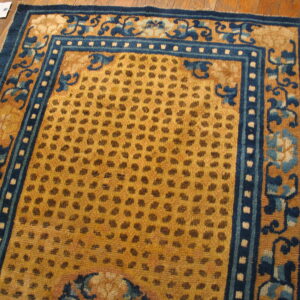 A gold rug with a spotted field and wide indigo blue scrolling floral borders rests on a wood floor.