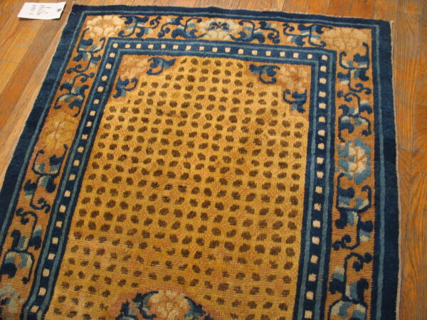 A gold rug with a spotted field and wide indigo blue scrolling floral borders rests on a wood floor.