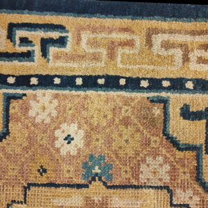 Close-up of a textured rug with gold, rust, and navy colors, featuring geometric borders and floral elements.