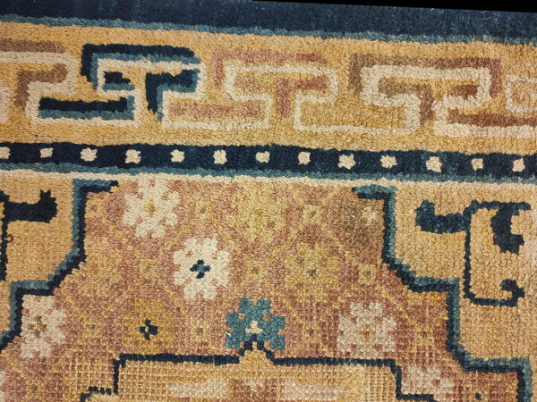 Close-up of a textured rug with gold, rust, and navy colors, featuring geometric borders and floral elements.