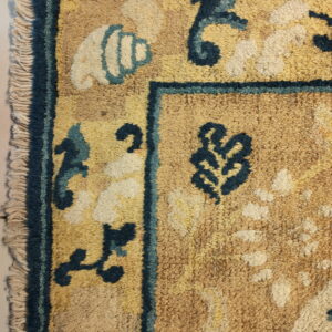 Detail shot of a shaggy tan rug featuring a dark blue border, cream fringe, and stylized teal and cream motifs.