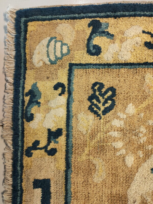 Detail shot of a shaggy tan rug featuring a dark blue border, cream fringe, and stylized teal and cream motifs.