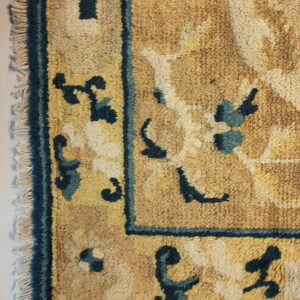 Textured tan and yellow rug corner with a decorative dark blue border and stylized cream floral motifs.