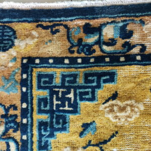 Medium-pile rug detail showing a golden field, navy key pattern, and teal stylized floral motifs with fringe.