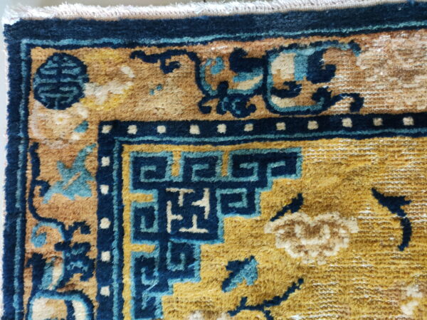 Medium-pile rug detail showing a golden field, navy key pattern, and teal stylized floral motifs with fringe.