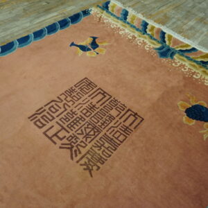 Dusty rose rug with brown block script, blue and yellow motifs, and complex cloud borders on distressed wood planks.