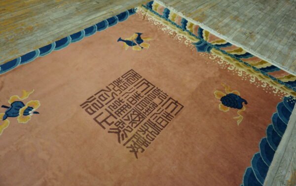 Dusty rose rug with brown block script, blue and yellow motifs, and complex cloud borders on distressed wood planks.