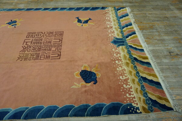 Pink rug featuring blue and yellow motifs, brown script, and multi-colored scalloped borders on worn wood planks.