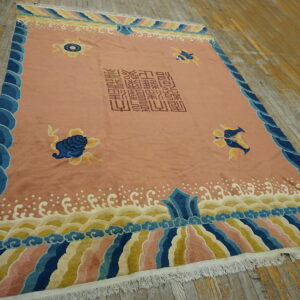 Pink low-pile rug with blue scalloped borders and central script lies on weathered wood floor.