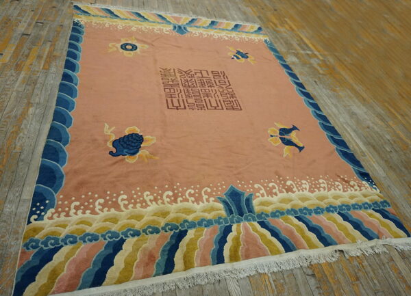 Pink low-pile rug with blue scalloped borders and central script lies on weathered wood floor.