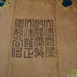 Detail of a reddish-tan rug showing dark brown Chinese characters and blue scalloped border design.
