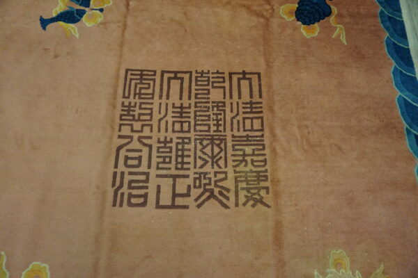 Detail of a reddish-tan rug showing dark brown chinese characters and blue scalloped border design.