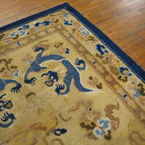 Close-up of a gold rug with blue dragon figures and a navy border on light brown hardwood floors.