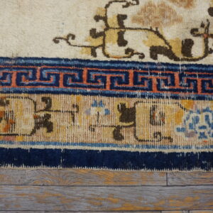 Cream rug with gold motifs and multiple blue, orange, and tan patterned borders lies on worn wood planks.
