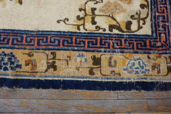 Cream rug with gold motifs and multiple blue, orange, and tan patterned borders lies on worn wood planks.