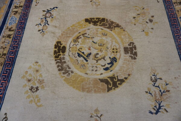Low-pile beige rug with gold central medallion, scattered florals, and a bold navy blue key pattern border.