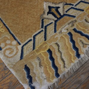 Geometric tan and blue rug edge showing medium pile texture over worn wooden floorboards.