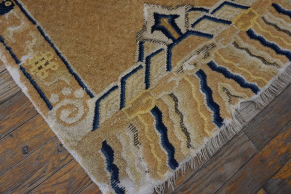Geometric tan and blue rug edge showing medium pile texture over worn wooden floorboards.