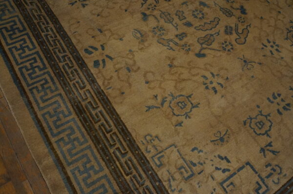 Tan rug with sparse blue floral patterns and a wide fretwork border on dark wood flooring.