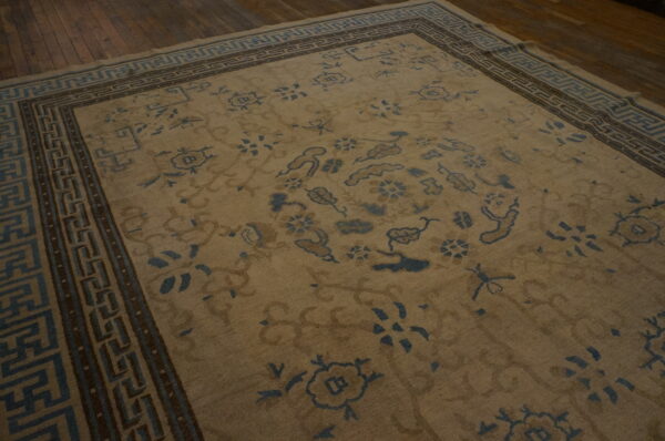 Low-pile tan rug featuring blue and brown floral patterns and key borders on dark hardwood.