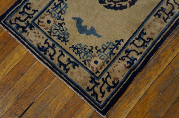 Corner of a tan rug with intricate blue and navy border designs on distressed hardwood planks.