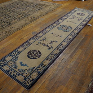 Beige runner rug with blue geometric designs and medallions on wood flooring next to a large dark patterned rug.