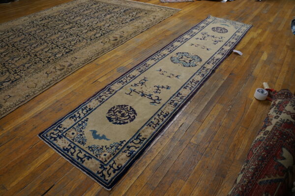 Beige runner rug with blue geometric designs and medallions on wood flooring next to a large dark patterned rug.