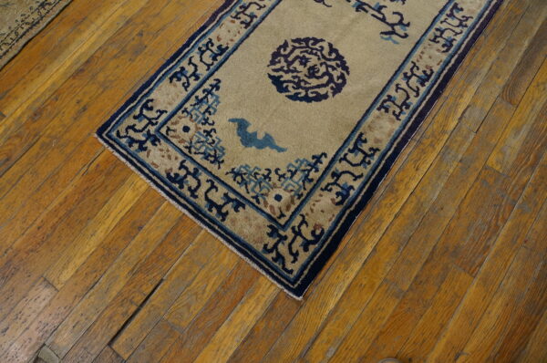 Tan rug with blue geometric borders and stylized patterns rests on distressed golden wood flooring.